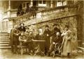 Gelatin silver photograph showing a seated group of men gathered around a small table in front of a building with a curved balcony and stone façade, early 20th century.