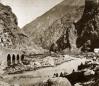Mountain gorge with a river flowing between steep rocky cliffs, featuring a stone bridge and a wooden bridge with people standing along the riverbank.