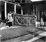 Two men holding a large carpet in a bazaar courtyard while other carpets are laid on the ground.