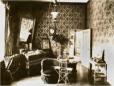Decorated interior room with patterned wallpaper, upholstered seating, mirror, framed portraits, small tables, and chandelier.