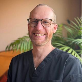 Forrest smiling in black scrubs, wearing glasses, with a tropical plant in the background
