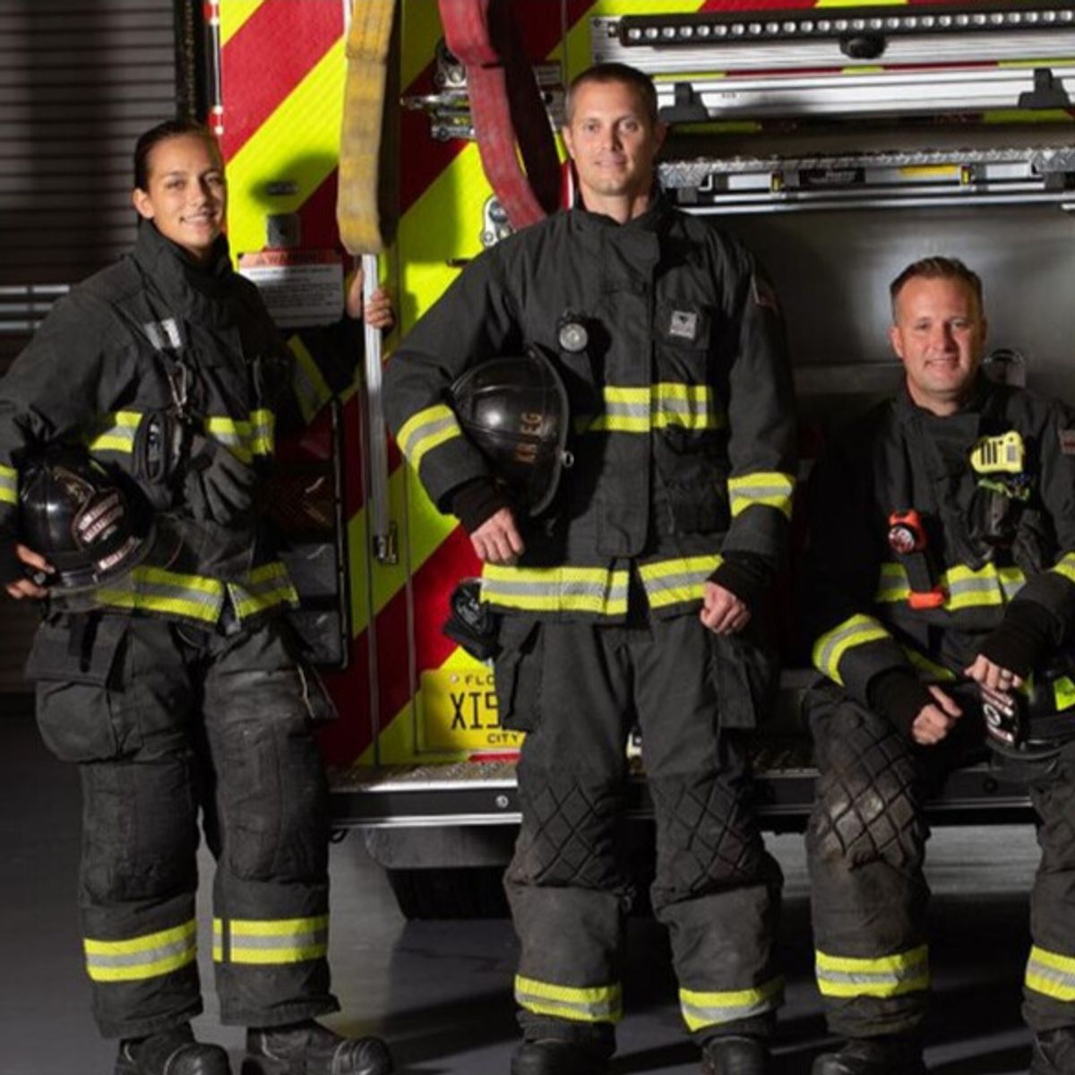 Three firefighters in gear holding helmets in front of firetruck