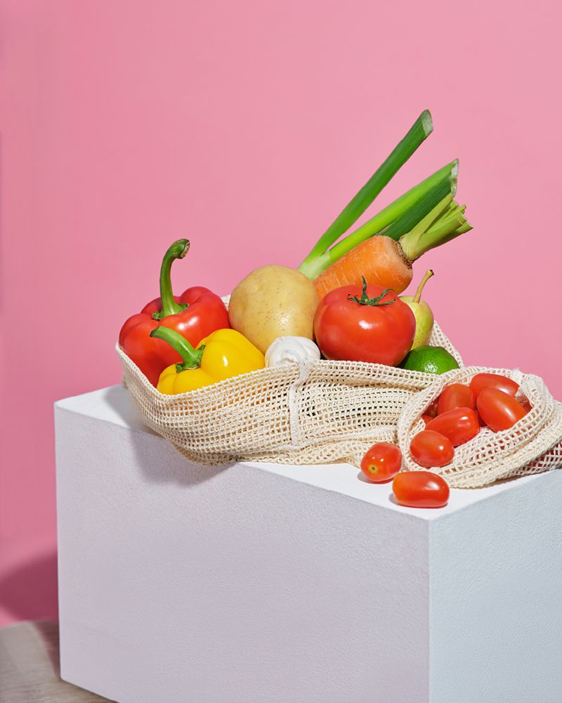 Fresh vegetables and produce arranged in reusable mesh shopping bags on a white plinth against a pink background, including red and yellow capsicums, tomato, carrot, potato, garlic and spring onions.