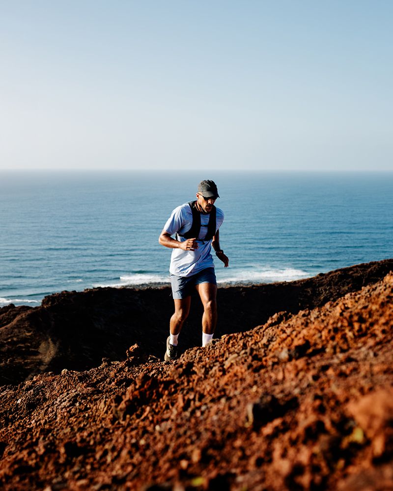 A man trail running along a rugged coastal clifftop with the ocean in the background under a clear blue sky.
