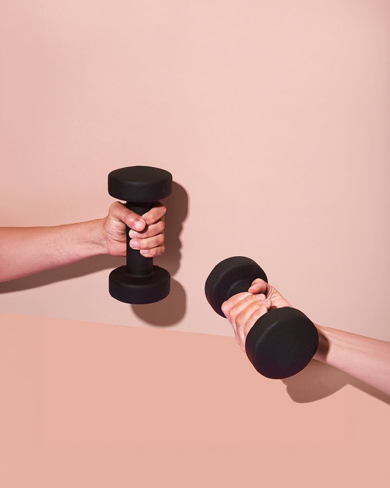 Two hands each gripping a black dumbbell, held up against a soft pink background