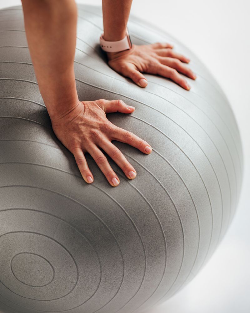 Two hands pressed flat on top of a large grey exercise ball, with a fitness tracker visible on one wrist.