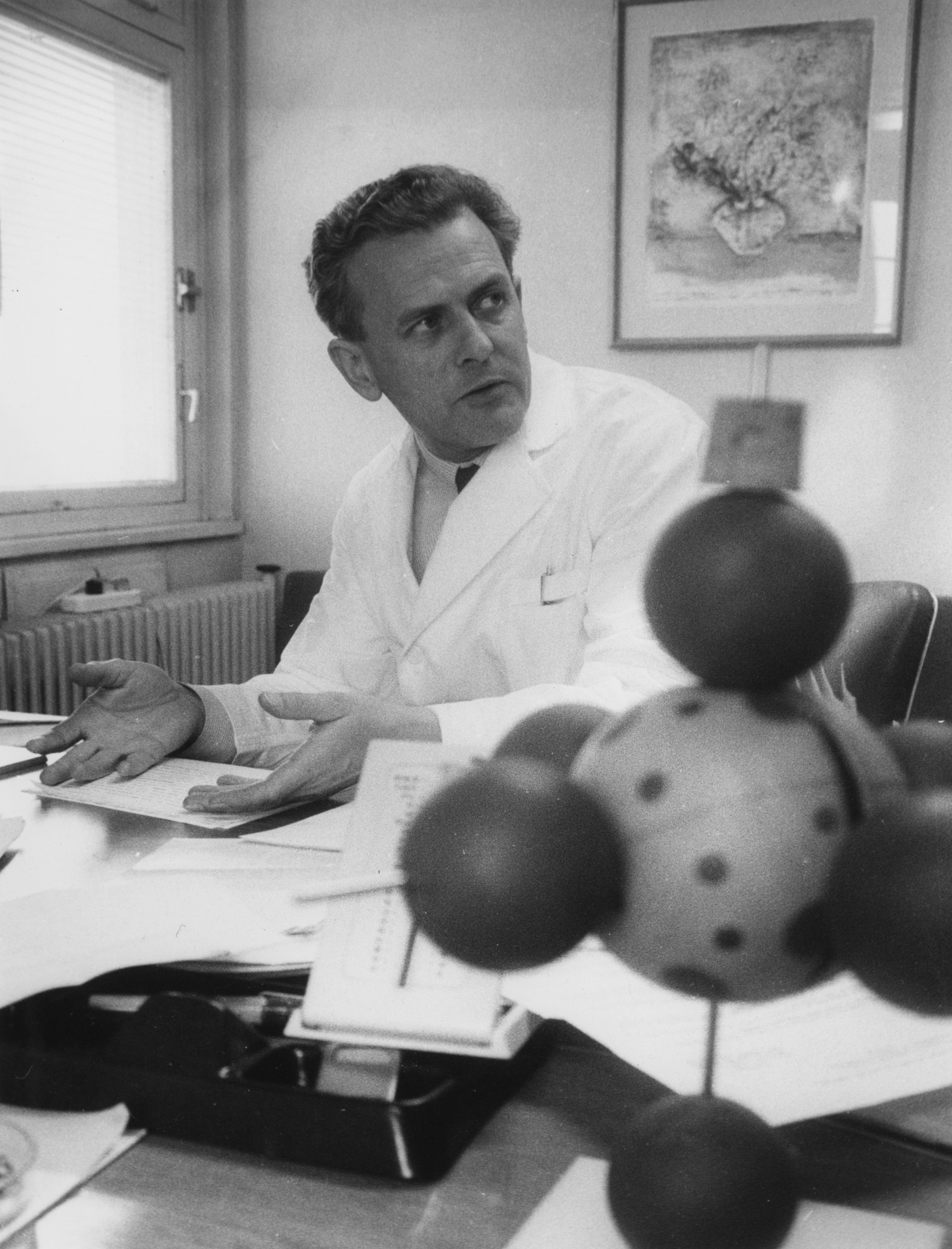 a man in a lab coat sits at a desk next to a model of a molecule