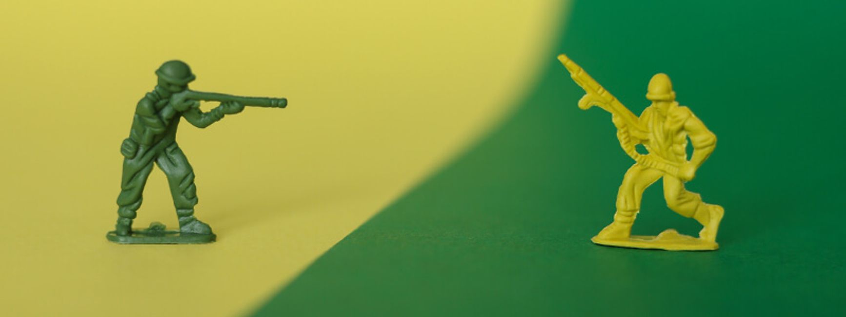 Two plastic army men toys pointing their guns at each other. They are two different colors.