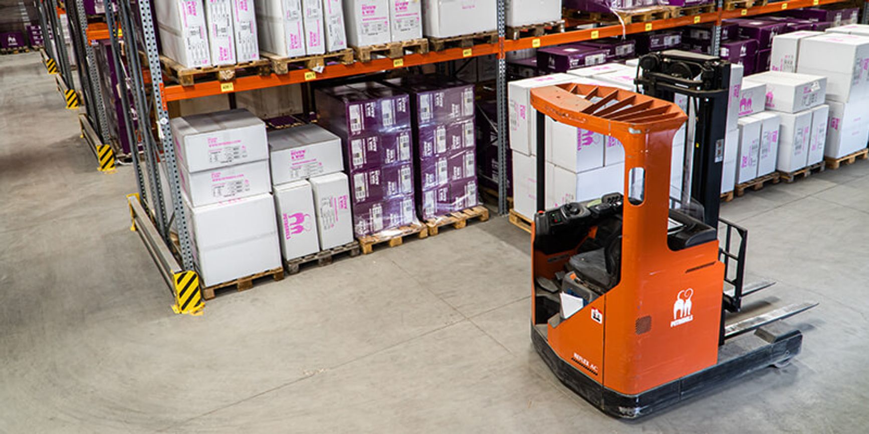 A forklift sits in the aisle of an organized warehouse full of shelves of boxes.