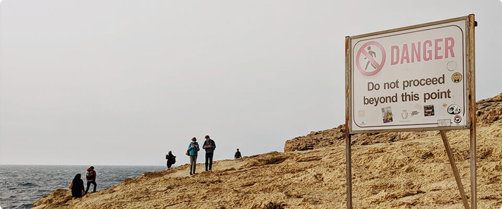 A sign on a rocky hill says "Danger: do not proceed beyond this point" and beyond the sign are 6 people exploring the hill.