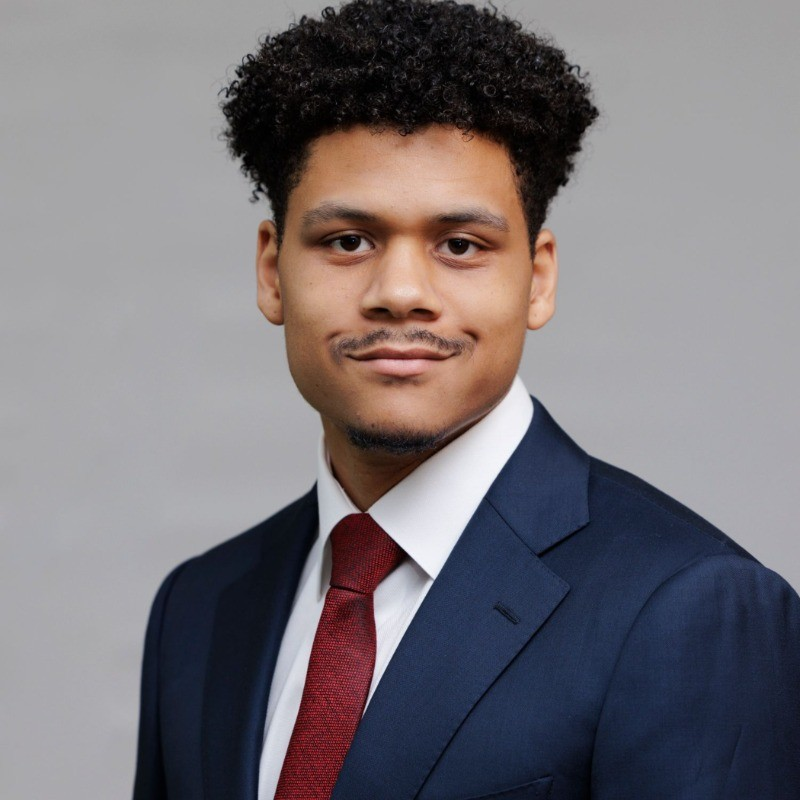A young man with curly hair and a mustache smiles in a navy suit and red tie against a gray background.