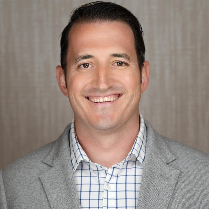 Headshot of a smiling man with dark hair, wearing a gray blazer and checkered shirt.