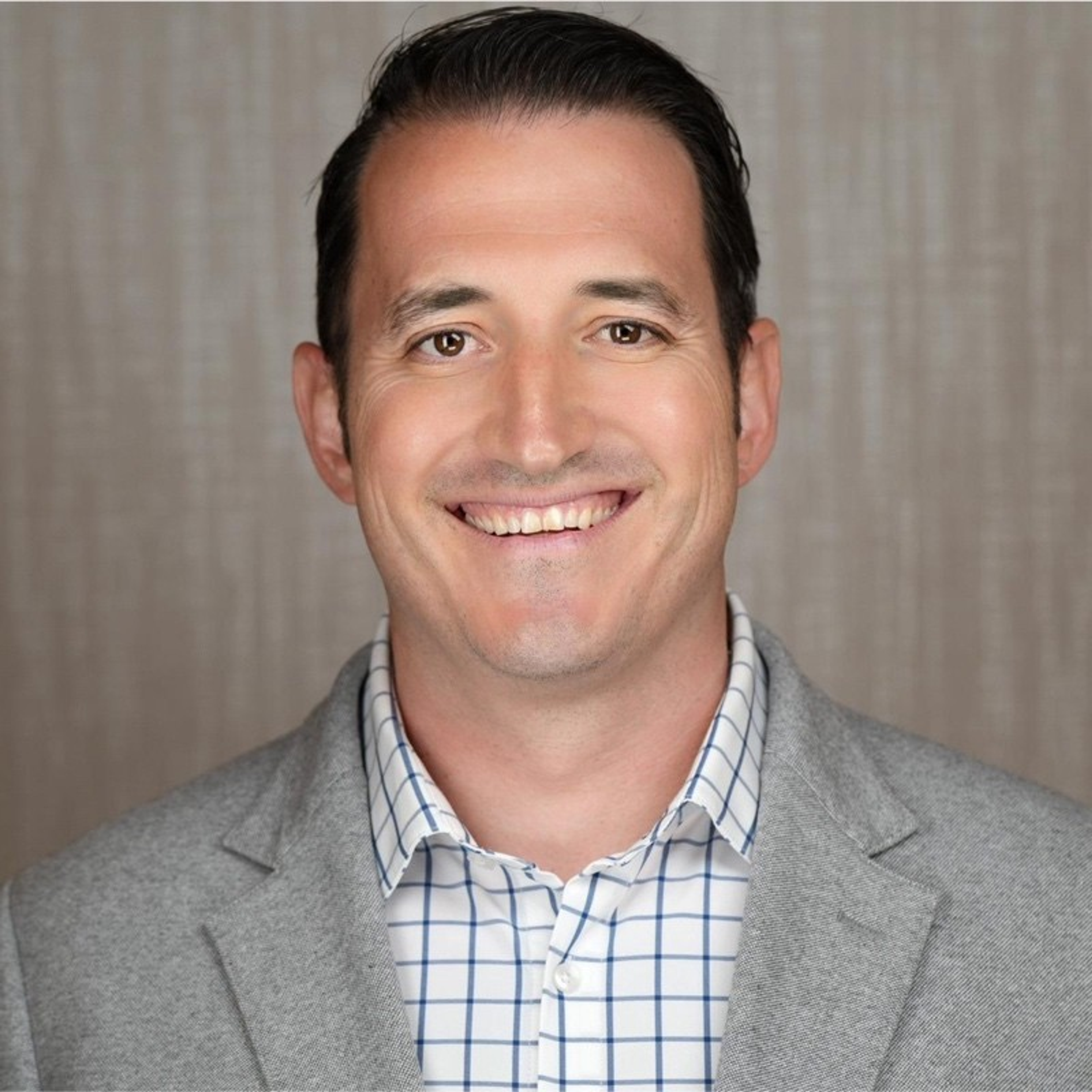 Headshot of a smiling man with dark hair, wearing a gray blazer and checkered shirt.