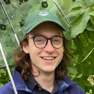 Smiling person with glasses, a green frog hat, and blue jacket, under a clear umbrella and green foliage.