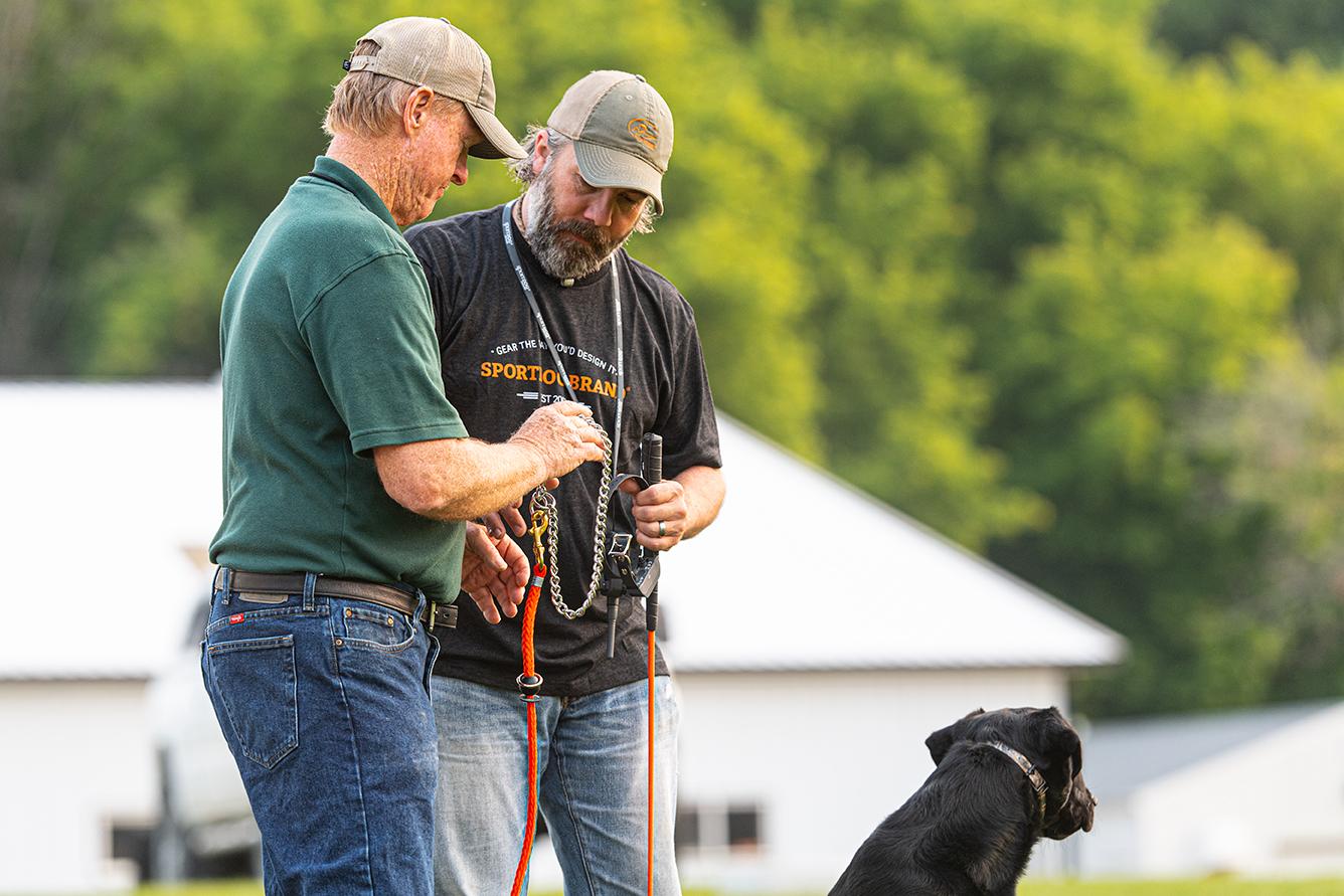Two men holding and talking about SportDOG training gear and a black lab sits next to them