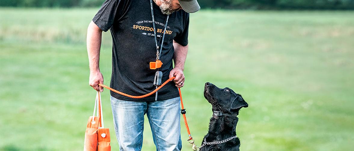 man looking at blck lab while holding leash and training dummies. Whistle and e-collar hanging from man's neck.