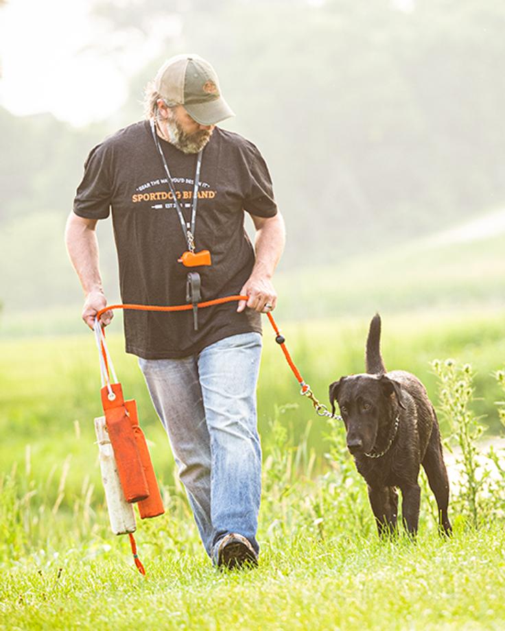 man walking black lab at heel while holding leash and canvas bumpers