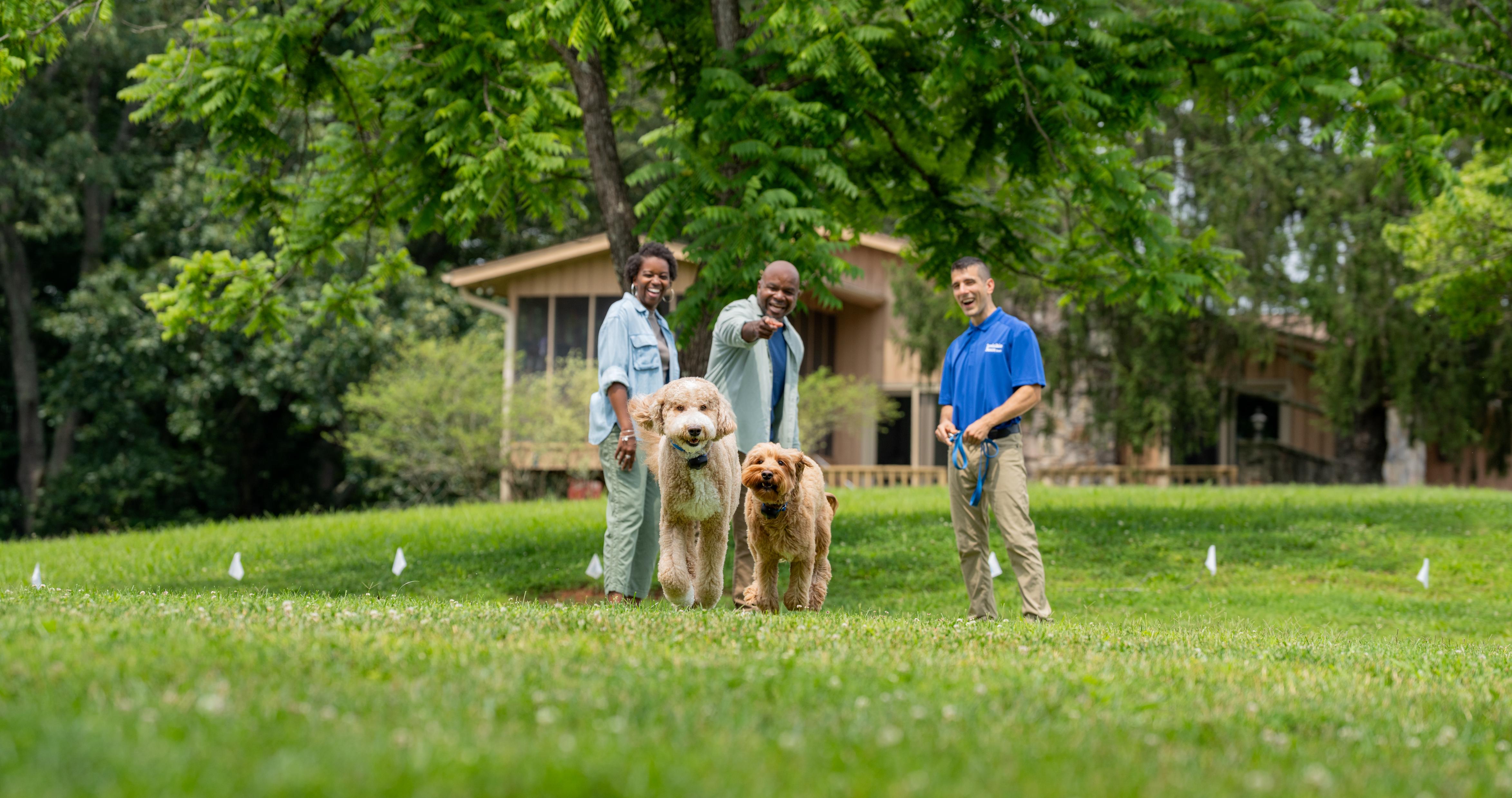 Two dogs running in yard that has a boundary plus in-ground system while owners watch excitedly alongside Invisible Fence Brand trainer.