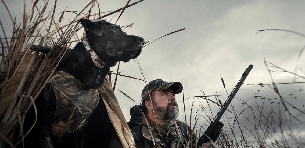 Dog wearing WetlandHunter collar next to man in camo holding gun