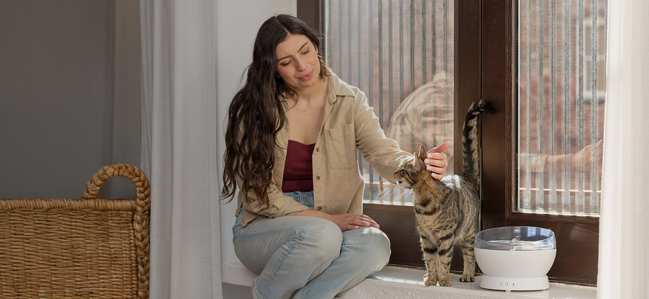 Woman sitting on windowsill petting cat next to white HydroSphere Cordless Pet Fountain