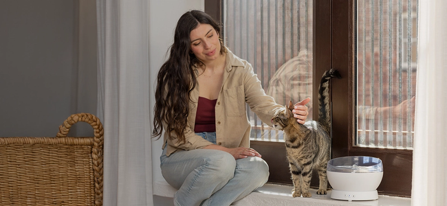Woman sitting on windowsill petting cat next to white HydroSphere Cordless Pet Fountain