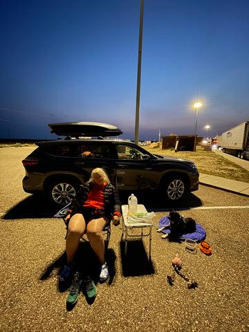 Jenny (founder of Unlikely Hikers) and Big Judy are resting on a camp chair and a dog bed in a parking lot.
