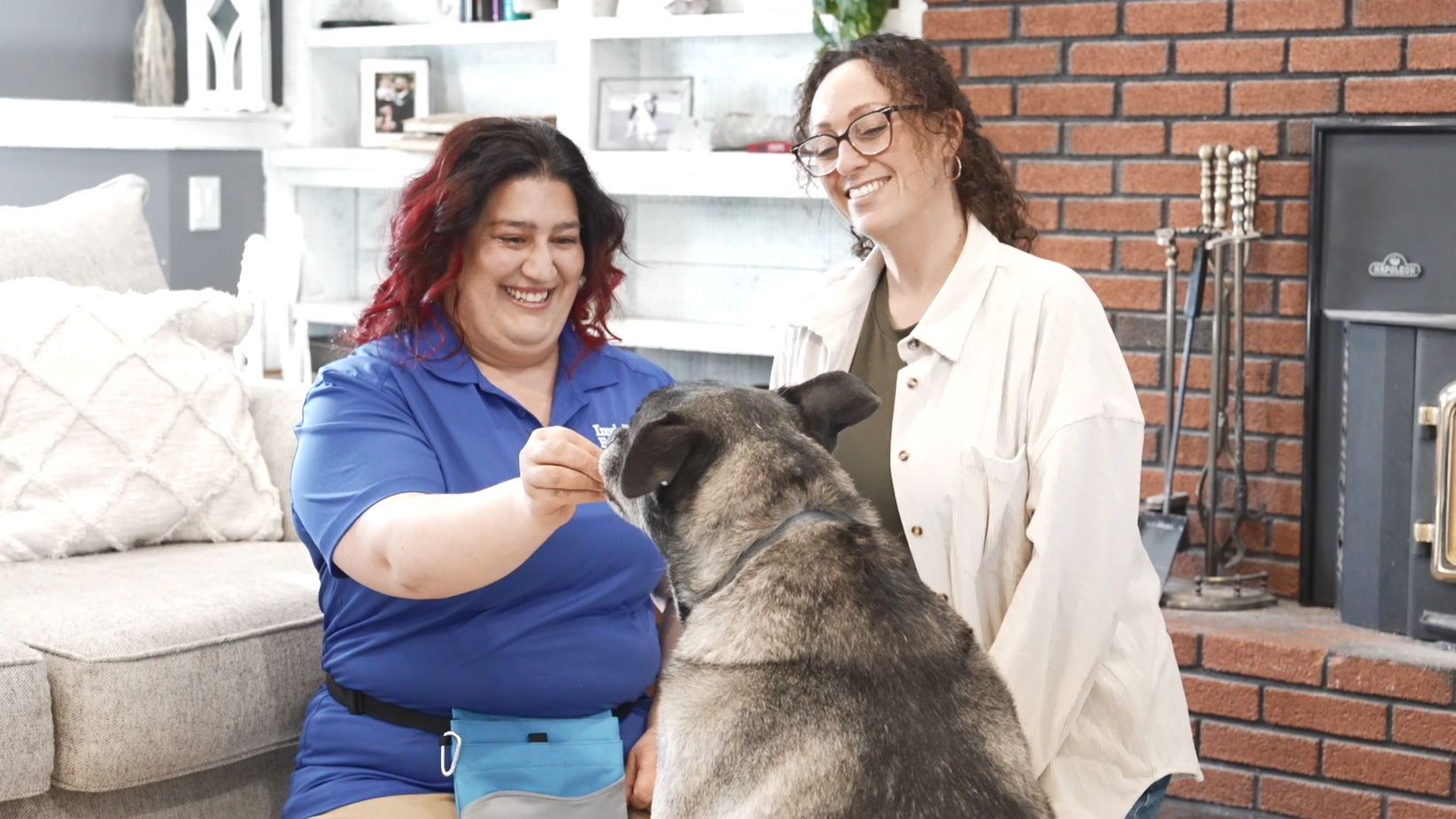 A client kneeling next to their Invisible Fence Manners Trainer who is luring a large grey dog into the sit position with a dog treat.