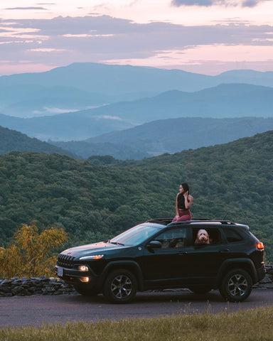 Sunshine sits in her parked car and looking at the sunset over the mountains. Sancho the goldendoodle is peeking his head out of the window.