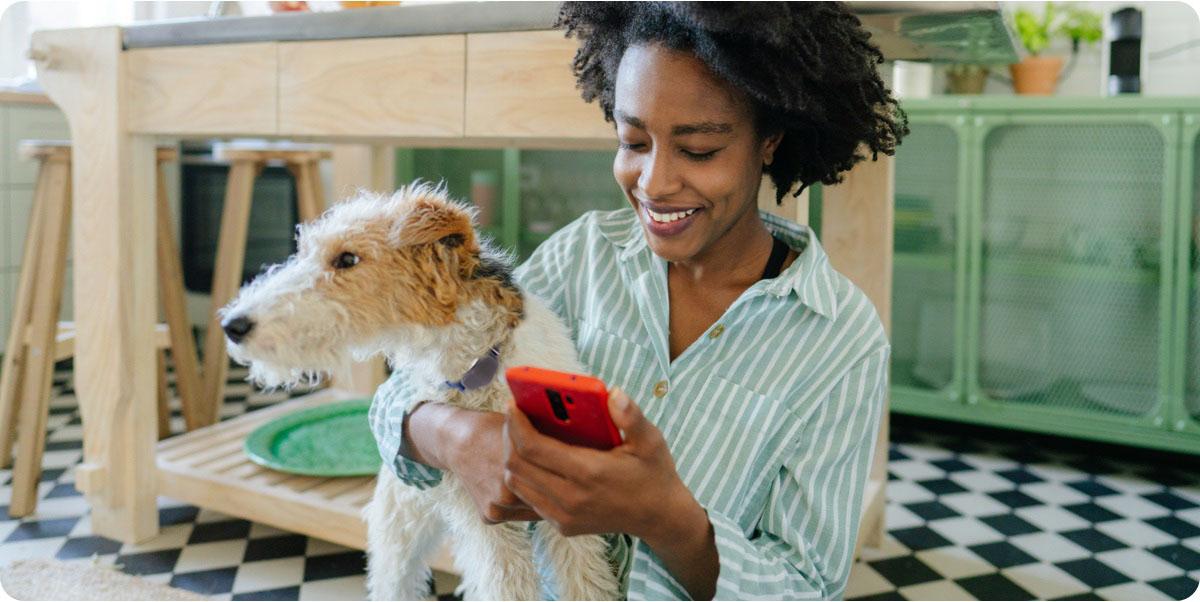 A woman sitting on the floor with her dog, holding a cell phone and smiling at the screen.