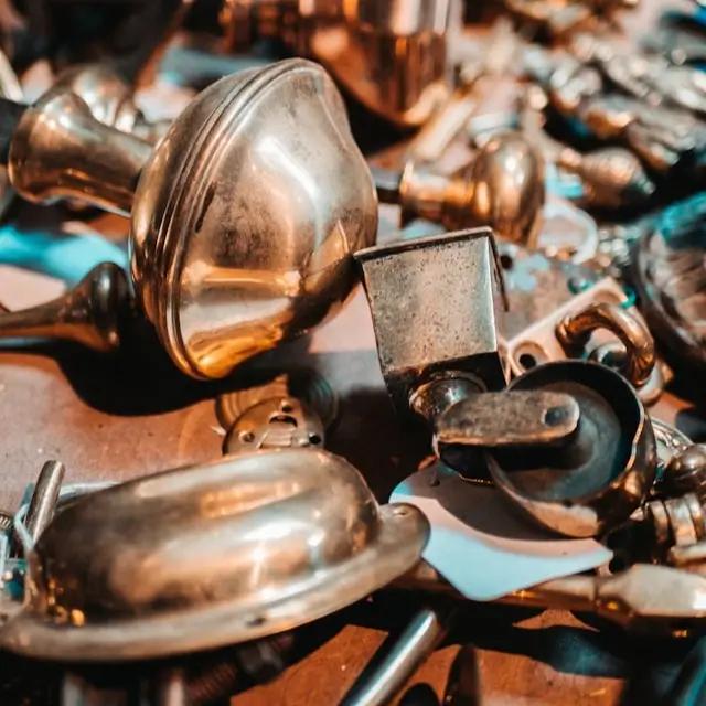 Close-up of a pile of assorted vintage brass doorknobs, handles, and other hardware components. The items are tarnished and worn, with some displaying a rich patina, indicating age and use. The mix of shapes and sizes creates a visually interesting collection, with the warm, reflective brass surfaces catching the light in various places.