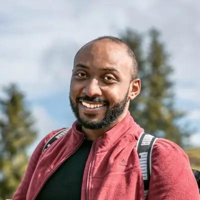 Sherif Negmeldin, Frontend Engineer for METYCLE; person wearing a black t-shirt and a red sweatshirt with the front zipper open, in a forest blurry background, smiling to the camera and with a beard