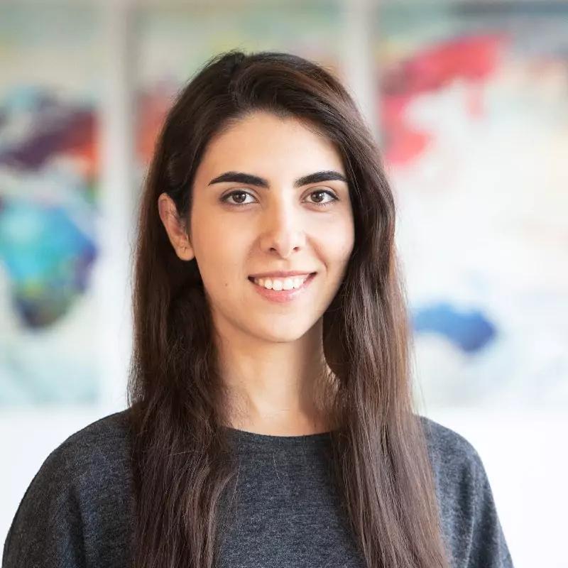 Nassim Raeesi, Senior Product Design for METYCLE; person wearing a dark gray shirt in front of a blurred colourful background, with long dark straight hair, smiling and looking directly to the camera
