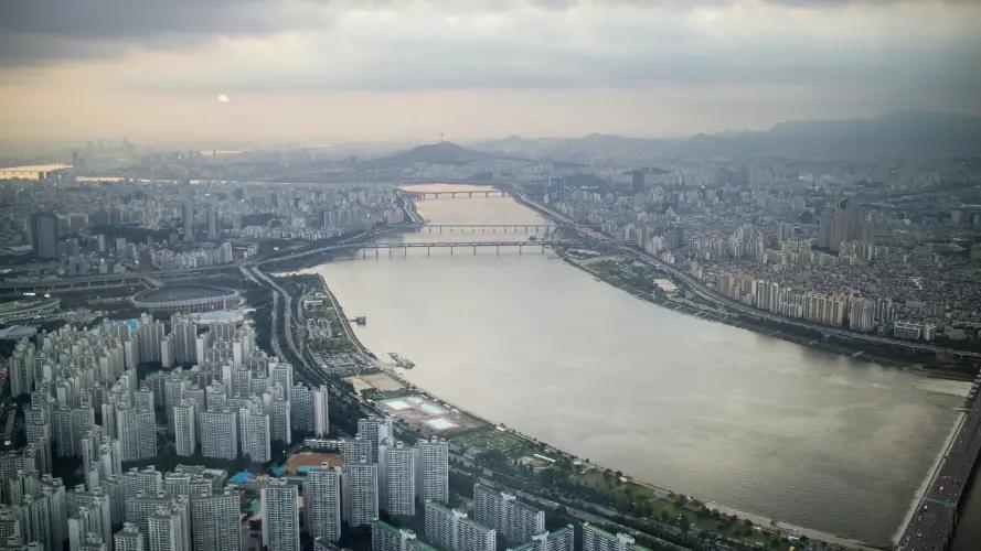 The image shows an expansive aerial view of a modern cityscape in South Korea dominated by a wide river running through the center. The city is densely packed with high-rise buildings, particularly clustered near the riverbanks. Multiple bridges span the river, connecting the two sides of the city. The sky is overcast, casting a muted, grayish light over the scene, giving it a calm, subdued atmosphere.