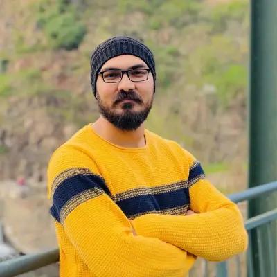 Usaid Sherwani, Working Student Frontend Engineer for METYCLE; person wearing a yellow sweater with a large dark and golden horizontal stripe in the chest, with the arms crossed, standing on a balcony, with a beard, glasses and a beanie