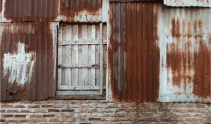 Exterior wall of a weathered building made of corrugated metal sheets and bricks. The metal sheets are rusted, with large patches of reddish-brown corrosion and areas of white paint peeling off. In the center, there is a small, boarded-up window with wooden planks.