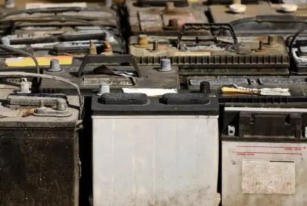 The image shows a collection of old, used car lead batteries lined up in rows. The batteries are various sizes and shapes, with many showing signs of wear, dirt, and corrosion. Some of the battery casings are discolored or stained, and the terminals are corroded or rusted.