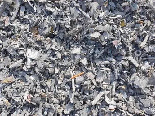 Large pile of shredded or broken pieces of grayish scrap metal. The fragments are irregular in shape and size, with a mix of jagged edges and more rounded pieces. The material appears densely packed, forming a textured surface. Among the mostly gray debris, a few small, colored items or fragments are visible, adding slight contrast to the otherwise monochromatic pile.