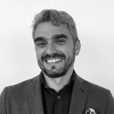 Aridany Ruano Freire, Software Architect for METYCLE via Edata Consulting; person wearing a dark gray suit and a black button down shirt in front of a light gray background, looking directly at the came and smiling, with wavy gray hair parted to the right side and a beard