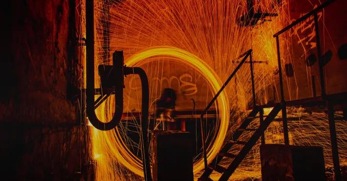 Industrial scene filled with vibrant, glowing orange sparks flying in all directions, from welding and grinding metal. At the center is a circular motion, creating a ring of bright light, while the background includes dark machinery and metal stairs. The intense sparks contrast against the dark surroundings, creating a dynamic and dramatic effect, highlighting the energy and heat of the industrial process taking place.