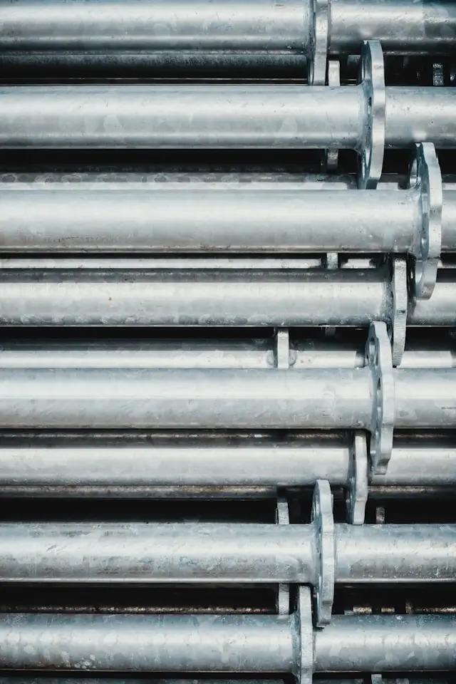 Stack of large, aluminum pipes arranged horizontally in a neat, parallel formation. The pipes are uniform in size, with smooth, shiny surfaces that reflect light. Each pipe has a flange at regular intervals, likely for connecting to other pipes or structures.