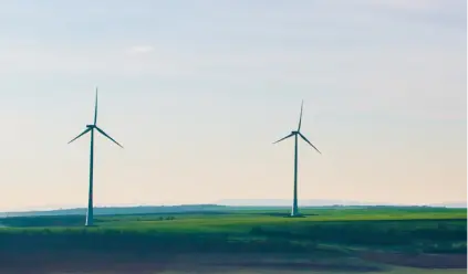 Landscape with two large wind turbines standing tall on a green field. The sky above is clear with a soft gradient from blue to light, indicating a calm day. The turbines' blades are still, suggesting little to no wind. The scene is peaceful, with the expansive field and distant horizon creating a sense of openness. The image highlights renewable energy through wind power, symbolizing a clean and sustainable approach to energy production.