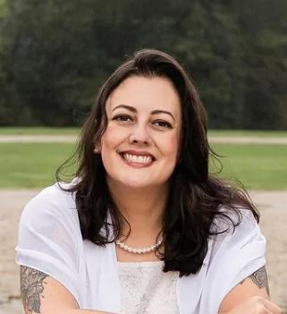 Camila Ribeiro, Head of Product, Engineering and Design for METYCLE; person wearing a white dress with tattoos in her arms, with a green field in the background, smiling and looking directly to the camera, with medium-length wavy dark hair parted to the right side