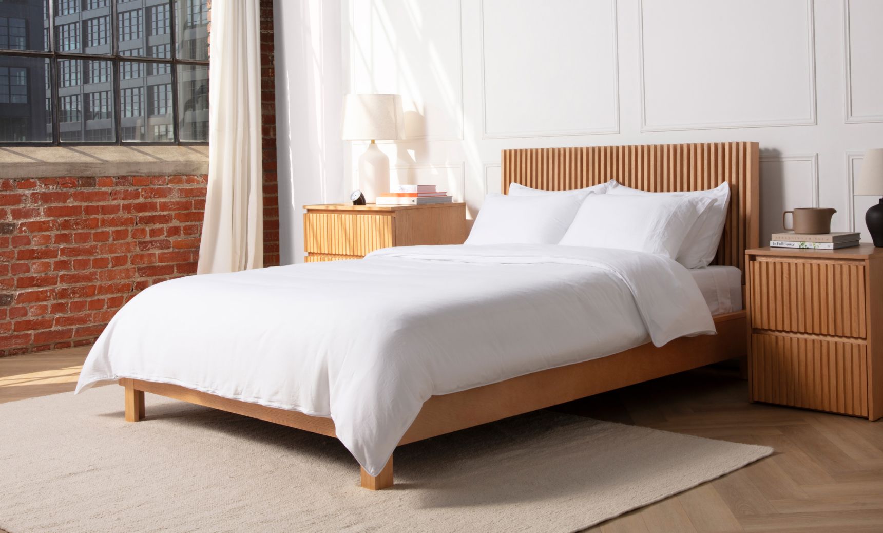 A fully dressed bed with a cozy white duvet and 4 pillows on a wooden frame, in a bedroom with a large window beside the bed