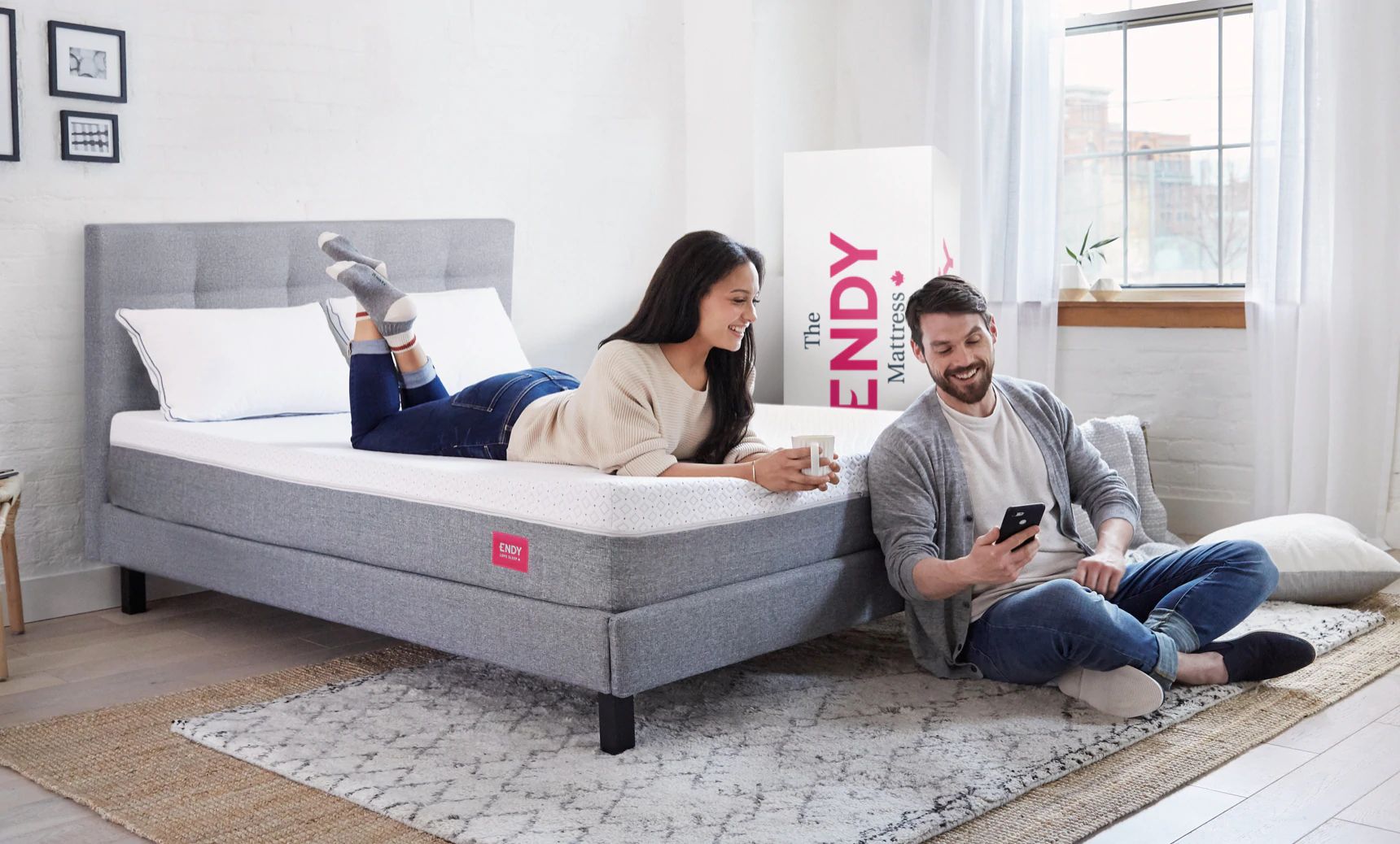 A woman laying on her stomach on a bare Endy mattress, looking at a phone held my a man sitting at the foot of the bed