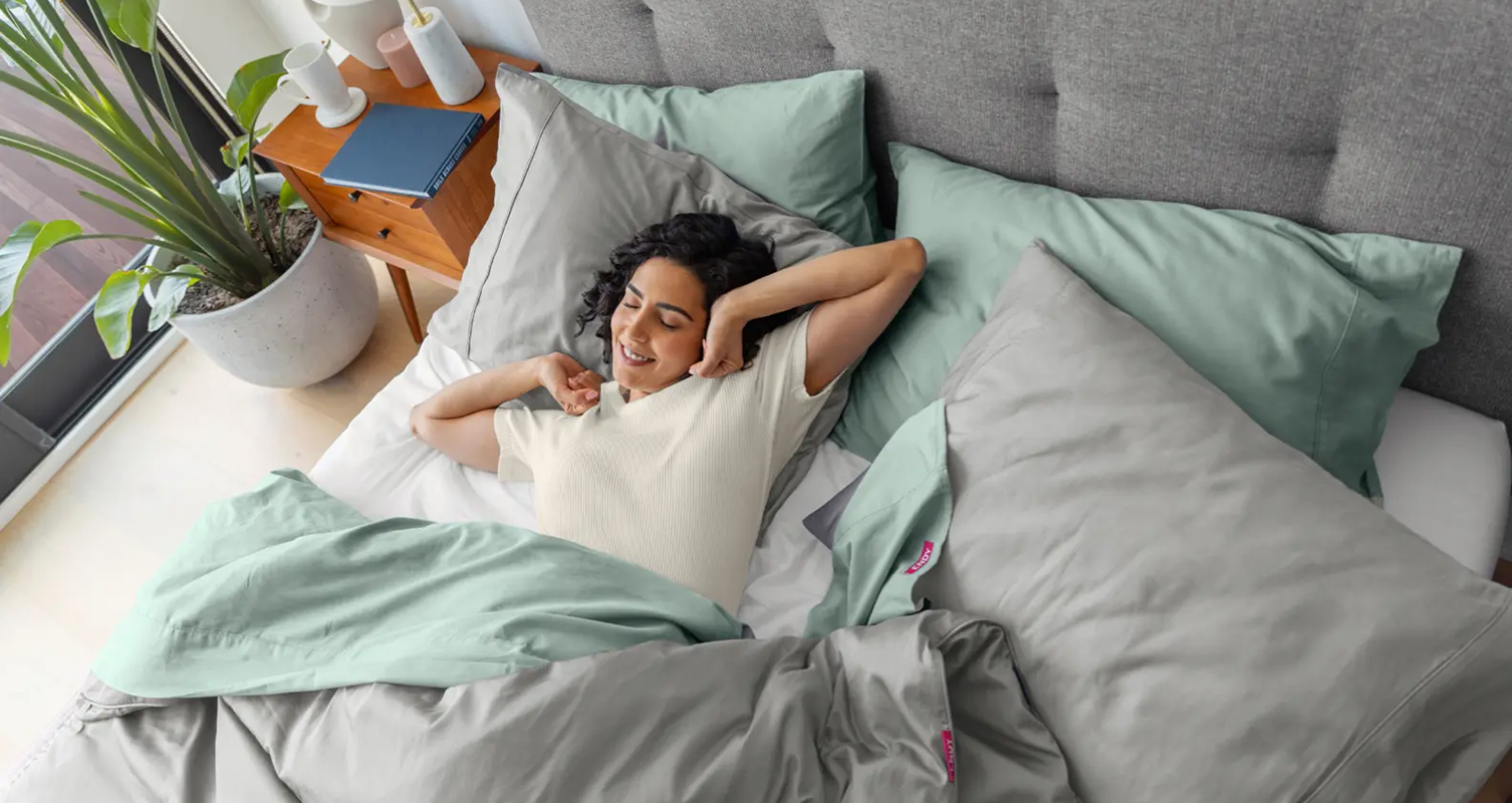 A woman stretching her arms in bed, wrapped in the Endy Organic Cotton Sheets in Soft Sage and the Endy Duvet and Organic Cotton Duvet Cover in Fog Grey.