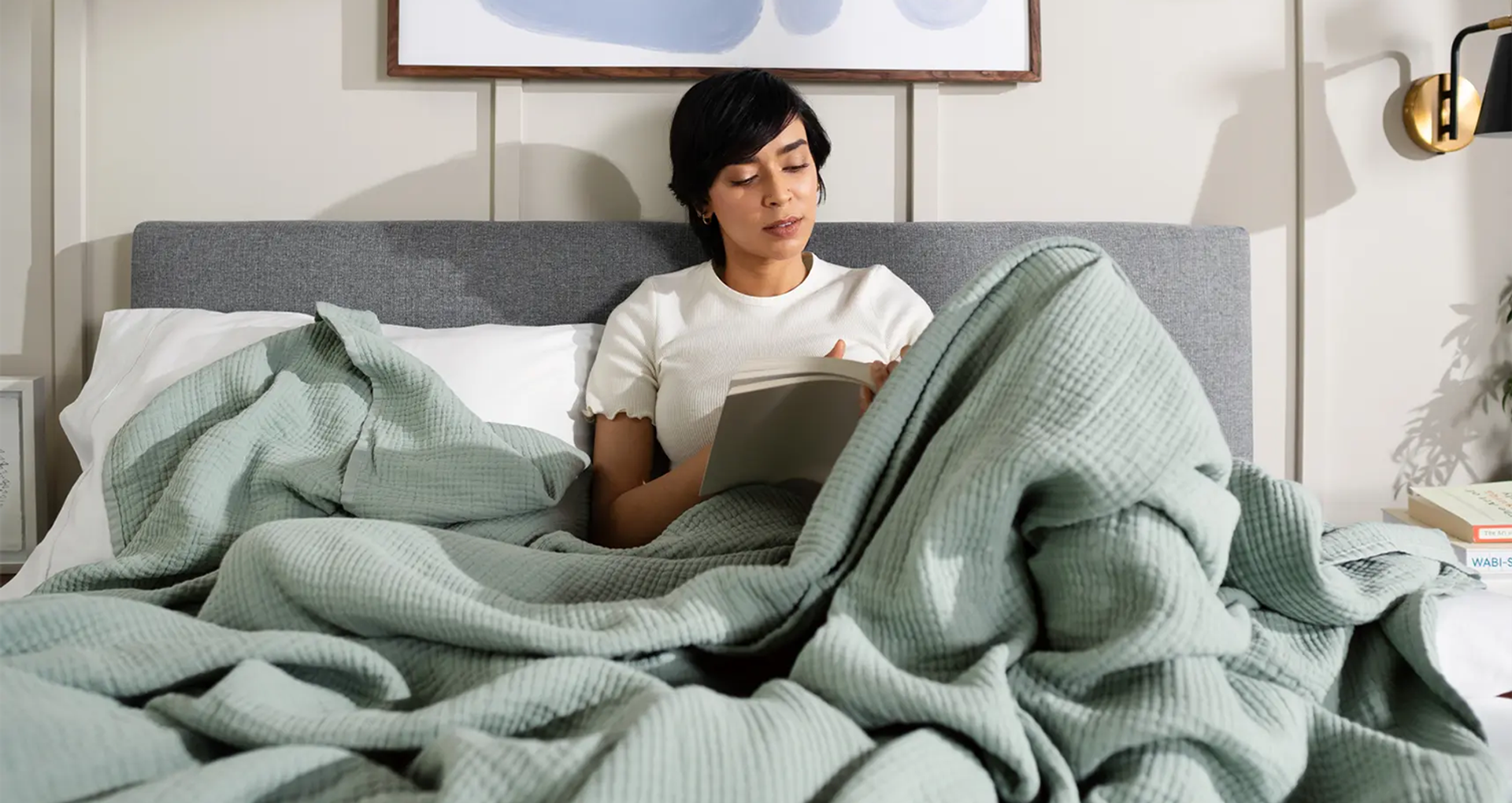 A woman sitting up in bed reading, wrapped in an Endy Muslin Blanket in Soft Sage