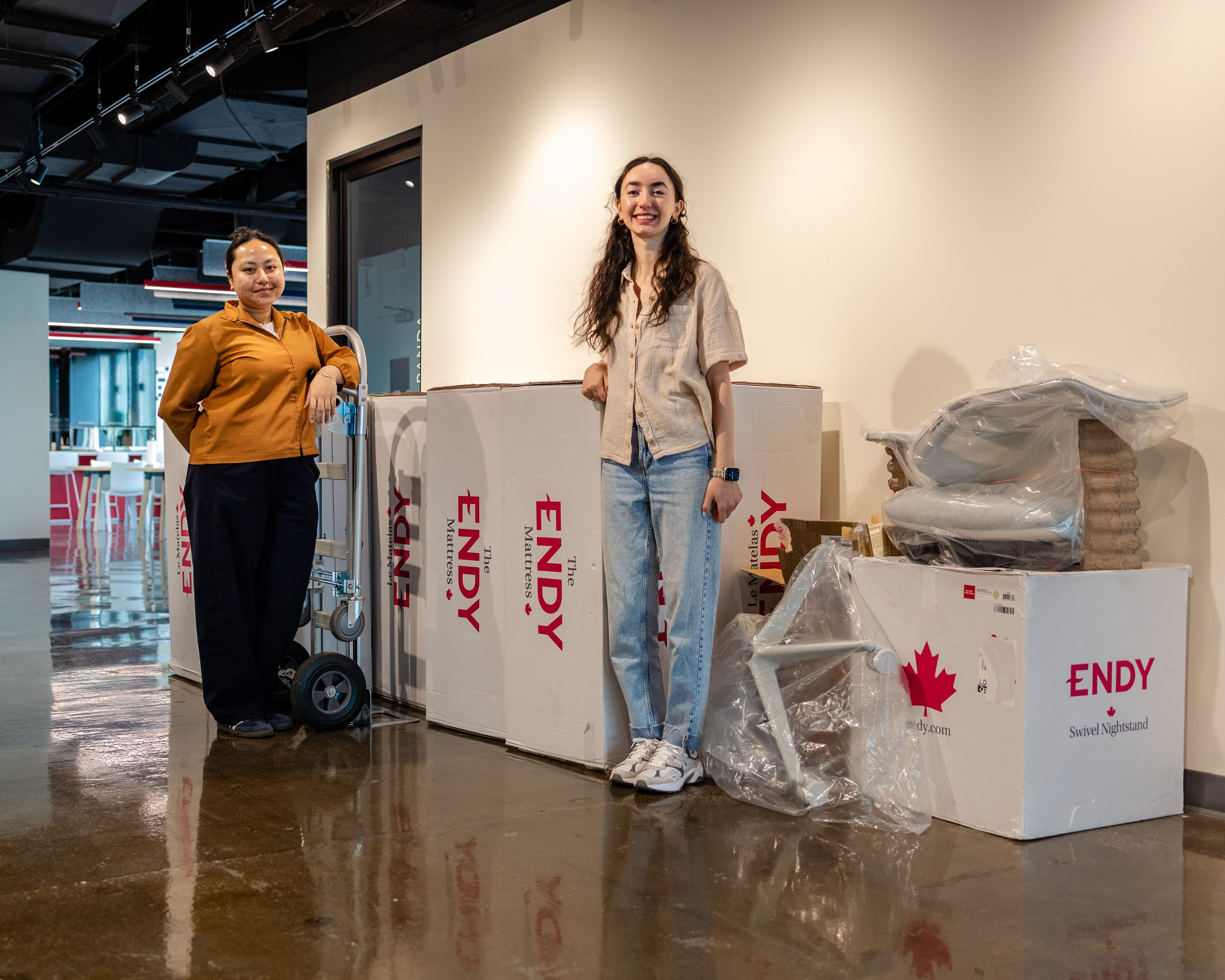 Two people standing in a hallway in front of several Endy Mattress boxes and a dolly.