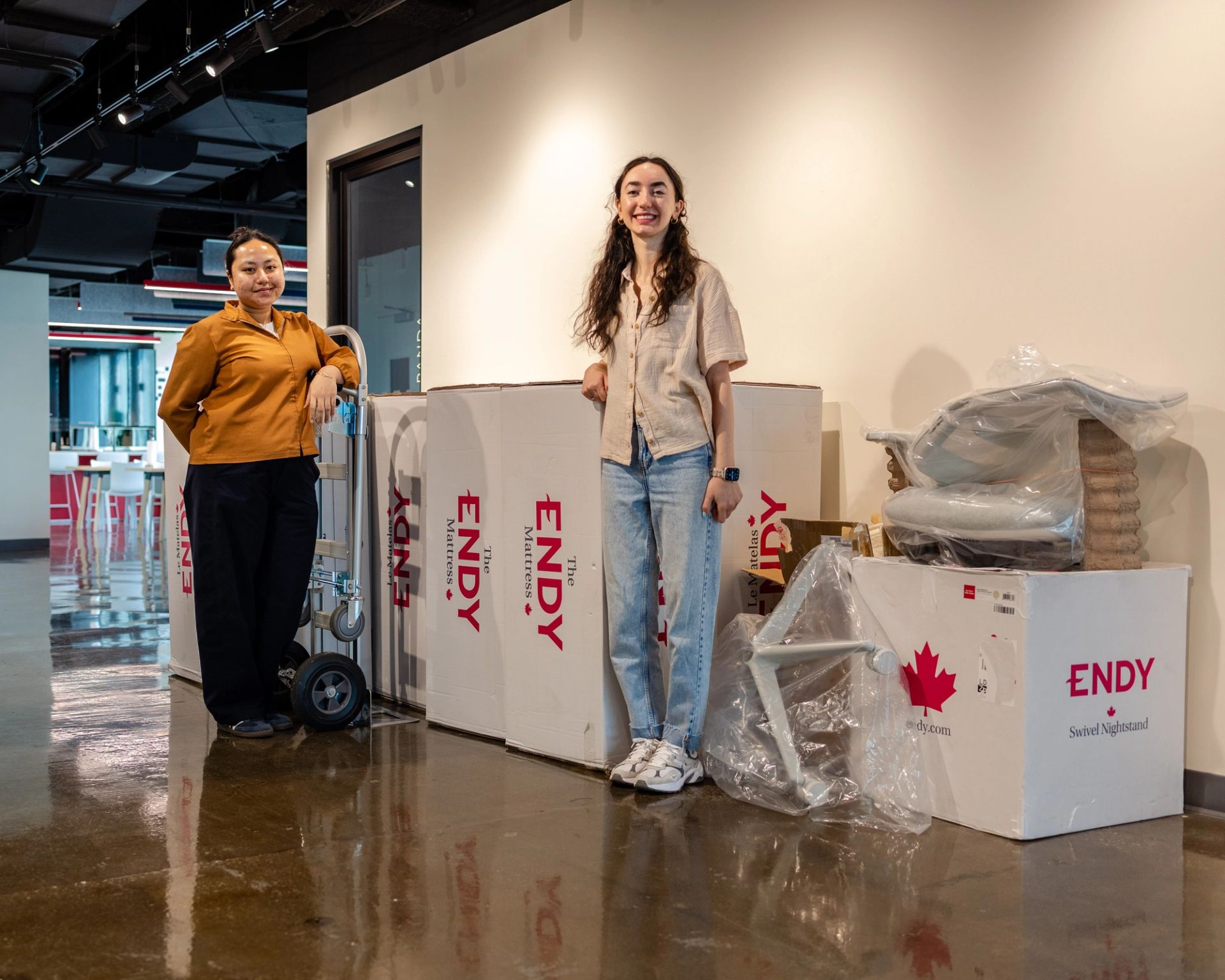 Two people standing in a hallway in front of several Endy Mattress boxes and a dolly.
