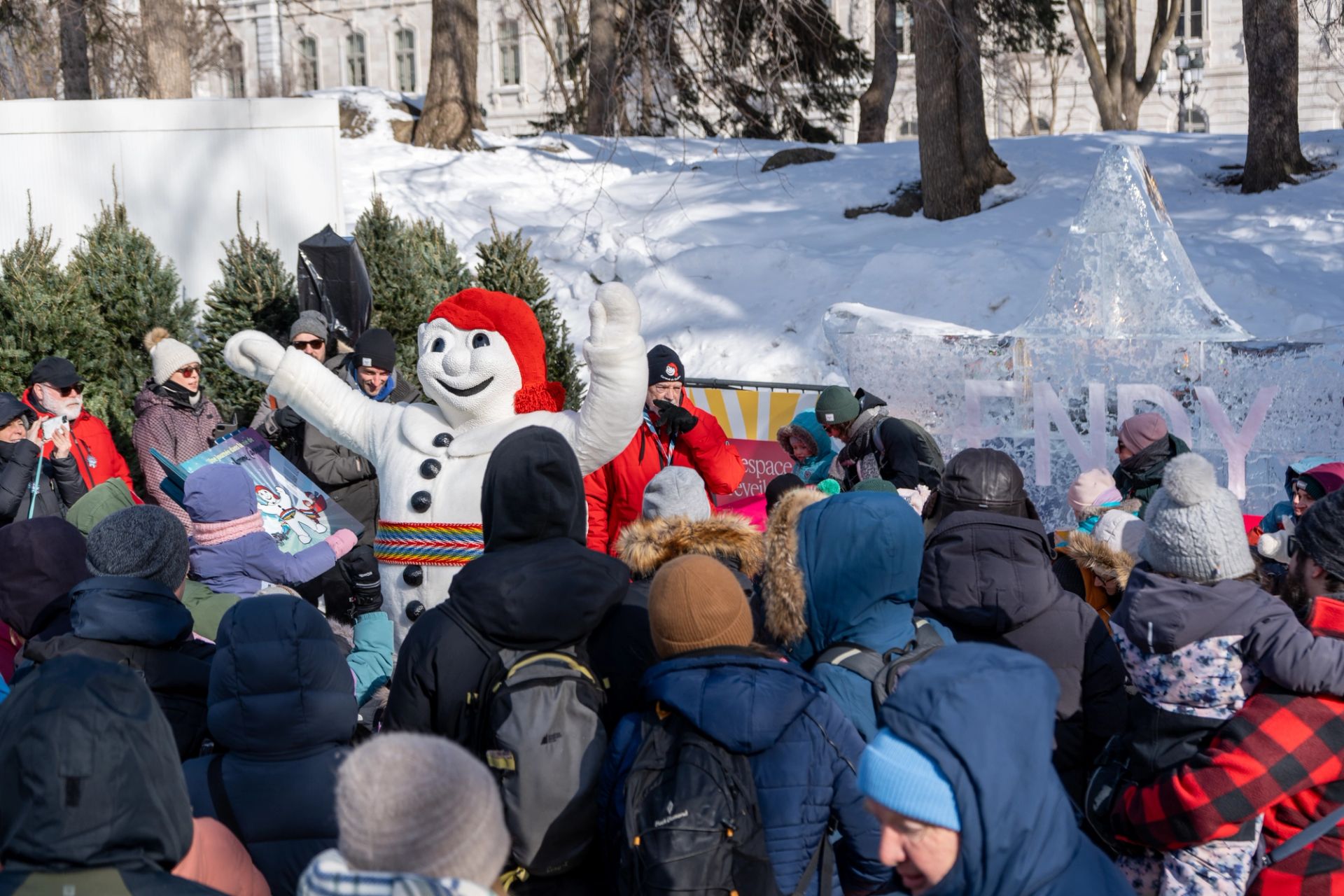 A large crowd of people dressed in winter gear gather around Bonhomme who is reading a book outside at Carnaval de Quebec