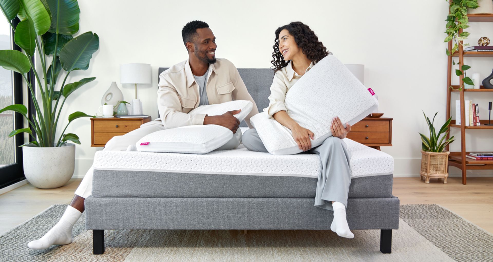 A couple sitting next to each other on the edge of an Endy Mattress, each hugging an Endy Customizable Pillow.